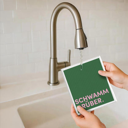 Schwammtuch mit grünem Text „SCHWAMM DRÜBER.“ in der Hand am Wasserhahn