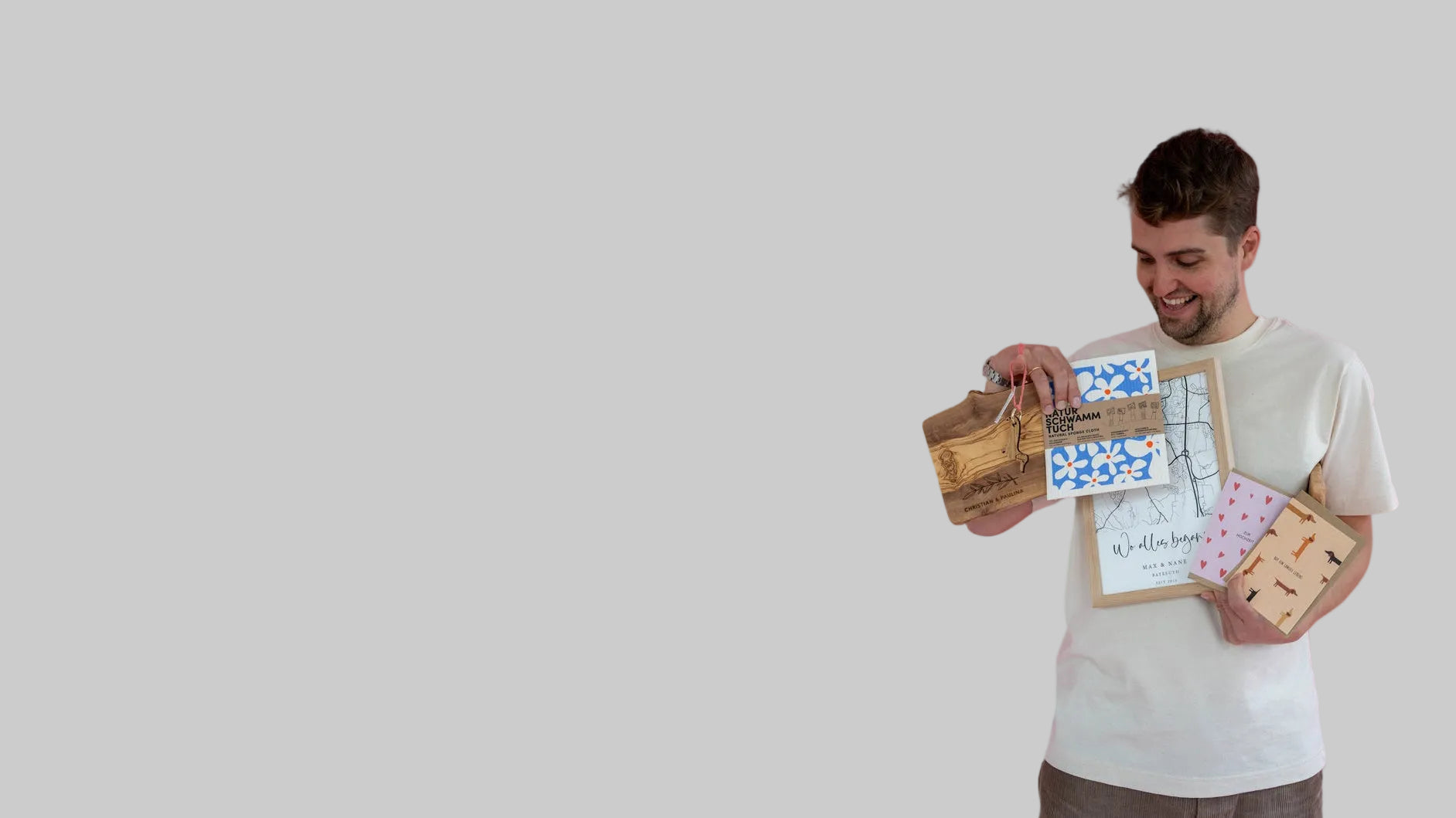 Das Bild zeigt eine Person, die ein personalisiertes Schneidebrett aus Olivenholz, ein Naturschwammtuch, ein Schlüsselanhänger aus Holz mit Dackel-Motiv und nachhaltige Karten in der Hand hält und von oben drauf schaut und lächelt.